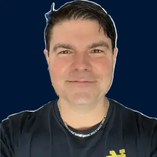 Smiling man with dark hair wears a navy blue shirt with a gold Notre Dame Music logo and silver necklace.