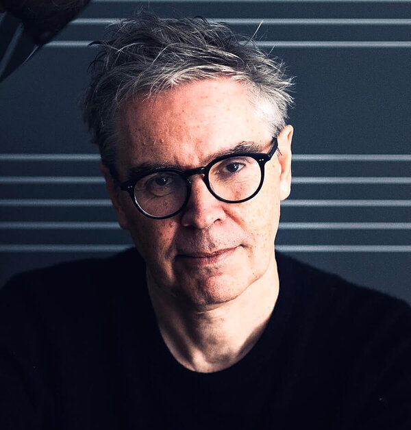 Man with messy gray hair, round black glasses, and a black shirt, looking directly at the camera against a dark striped background.